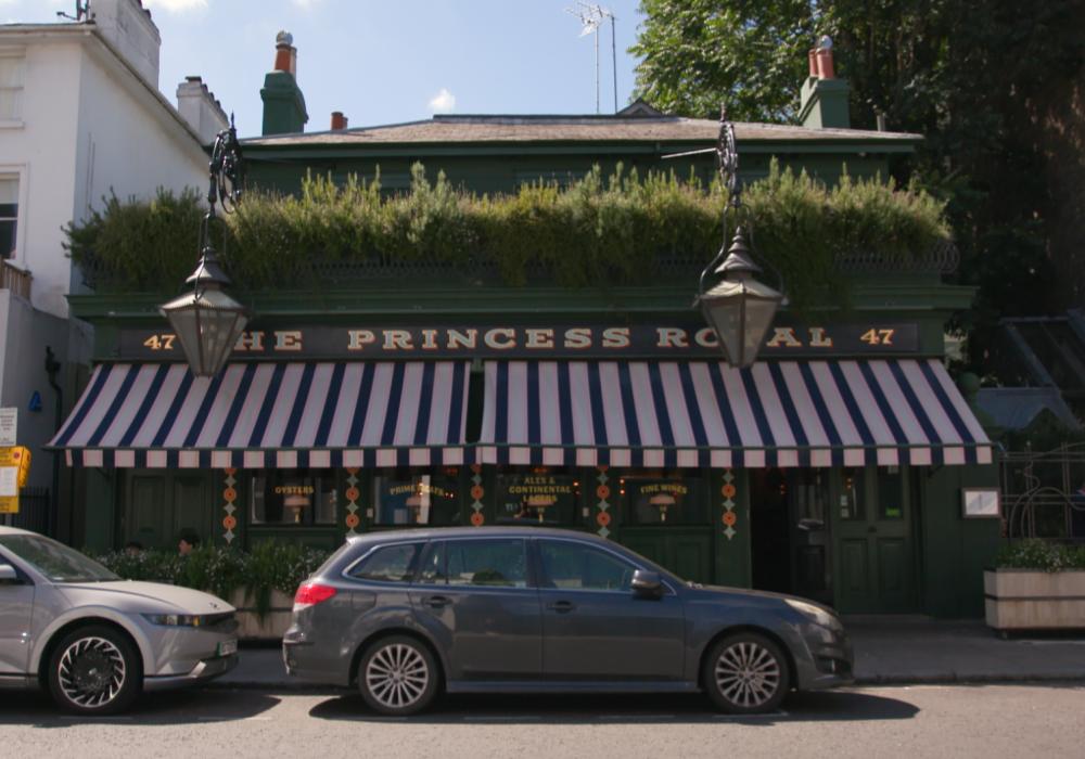 The exterior of a London pub with red and white awning outside and greenery on the front of the building