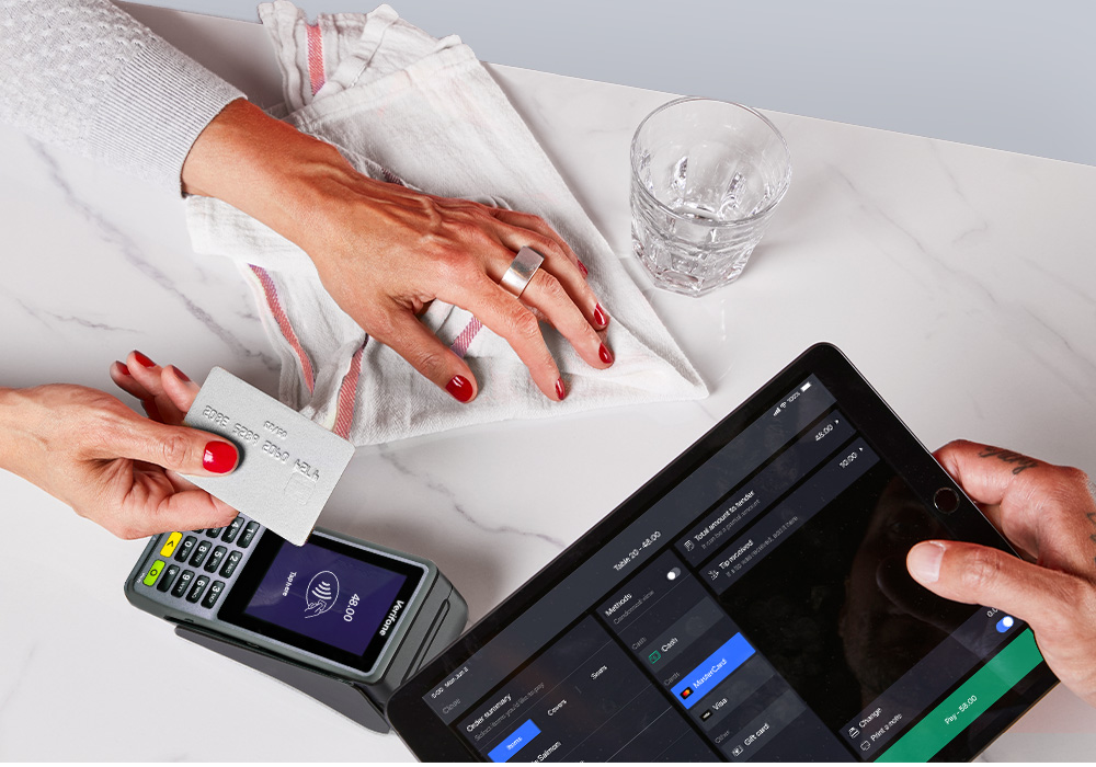 A person holding a silver credit card and is about to tap it on a payment terminal. In the image is a iPad displaying what the customer owes