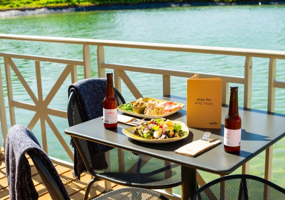 A table and chairs set by the river. The table is laid with two plates of food and two bottles of beer