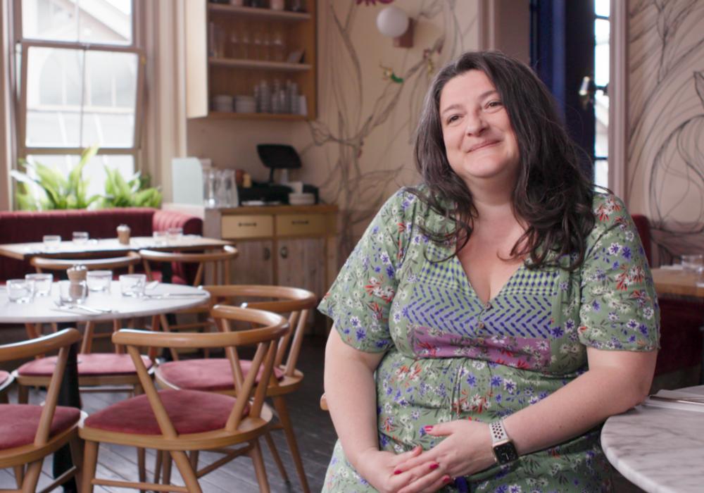 A photo of a woman sat smiling at the camera with tables and chairs in the background