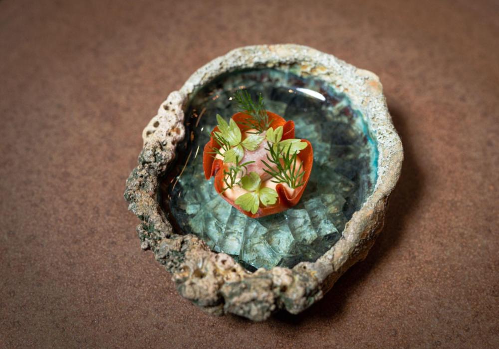 A small rugged bowl filled with a small red tart case with eel and green leaves in the centre