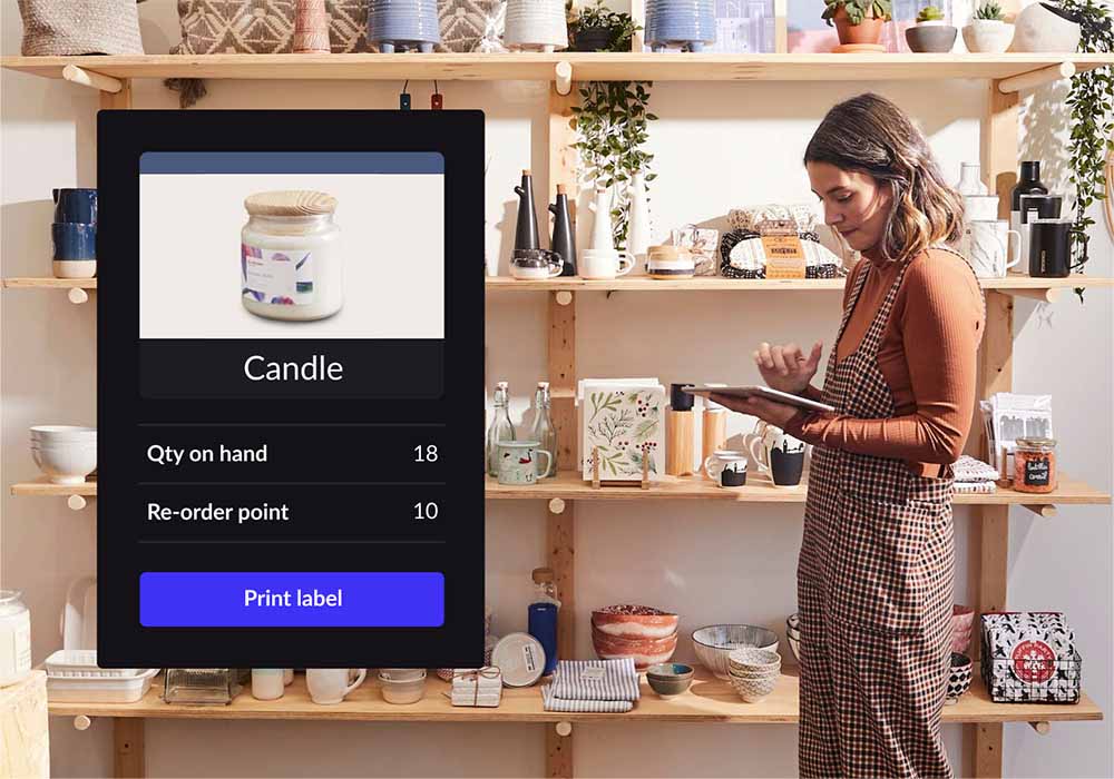 Scented candle shown on Lightspeed Retail's inventory management interface. Quantity on hand, re-order point and product categories are shown below it. Retailer using iPad in shop in background.