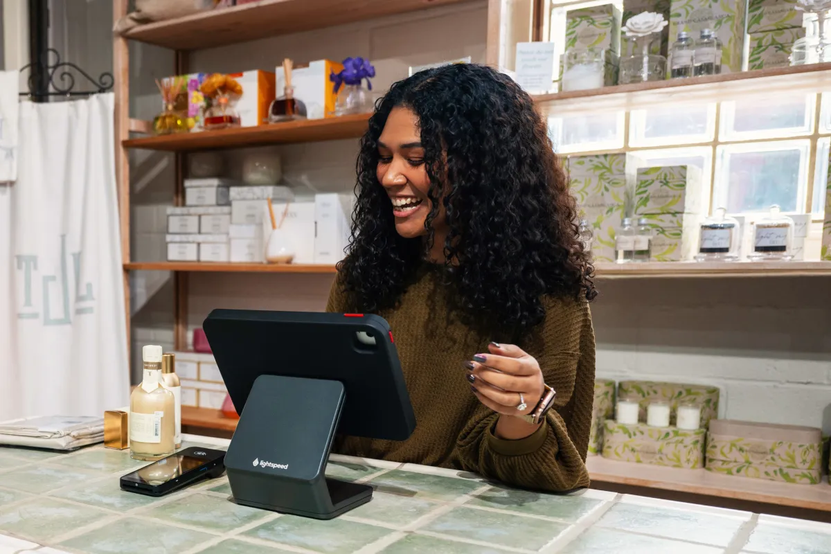 A woman grins behind the counter of a store while looking at a Lightspeed point of sale screen.