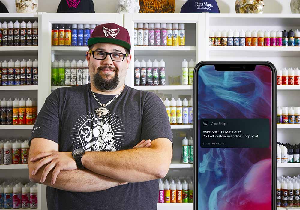 iPhone showing loyalty notification, retailer smiling in front of shelves stocked with vape pens