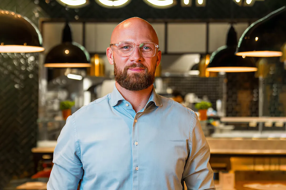 A man poses in front of a kitchen at a restaurant.