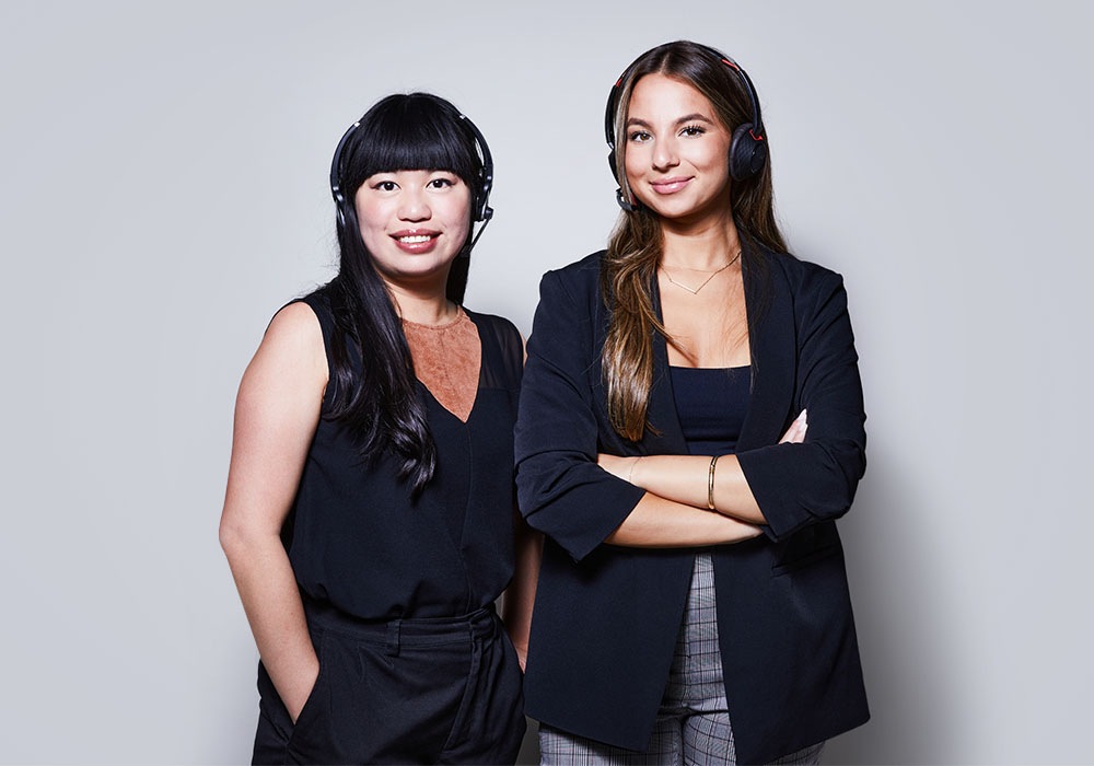 Image of two women with headsets on, wearing black clothing and smiling at the camera
